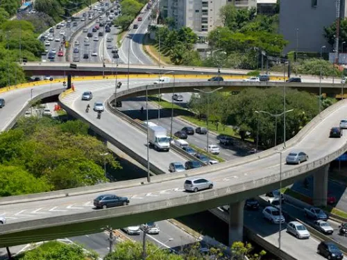 Vias elevadas com tráfego de carros, rodeadas por vegetação e prédios ao fundo.